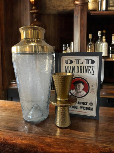 An upscale bar essentials set for the modern gentleman, including a gold cocktail shaker and matching jigger, rests on a wooden bar counter. Behind them is the book 