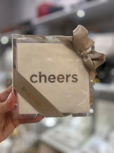 Load image into Gallery viewer, A hand holds a Cocktail Napkin Acrylic Tray Set - Year Round Entertaining, featuring the word &quot;cheers&quot; printed in bold. They are beautifully encased in a clear plastic box tied with a gold ribbon, making it an ideal hostess gift. A diagonal label indicates the &quot;lined design.&quot; In the softly blurred background, shelves brimming with various items can be seen.
