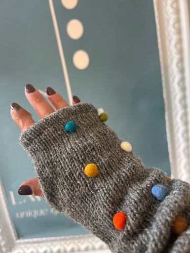 A hand with dark nail polish is wearing the Pom Pom Wool Fingerless Gloves, which are gray knit gloves adorned with vibrant round pom-poms. Made from sheep wool, these gloves expertly blend warmth and style. The background features a blue color with a white and gold frame, highlighting an abstract design with dots and partially visible text.