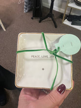 Load image into Gallery viewer, A hand with dark red nail polish holds the Holiday Soap in a Dish, a small square ceramic piece adorned with gold edges that exudes holiday cheer. Wrapped in a green ribbon and featuring a round light green tag, the dish displays &quot;PEACE. LOVE. JOY&quot; above a small star. In the background, carpeted floors and shelves are filled with festive delights.
