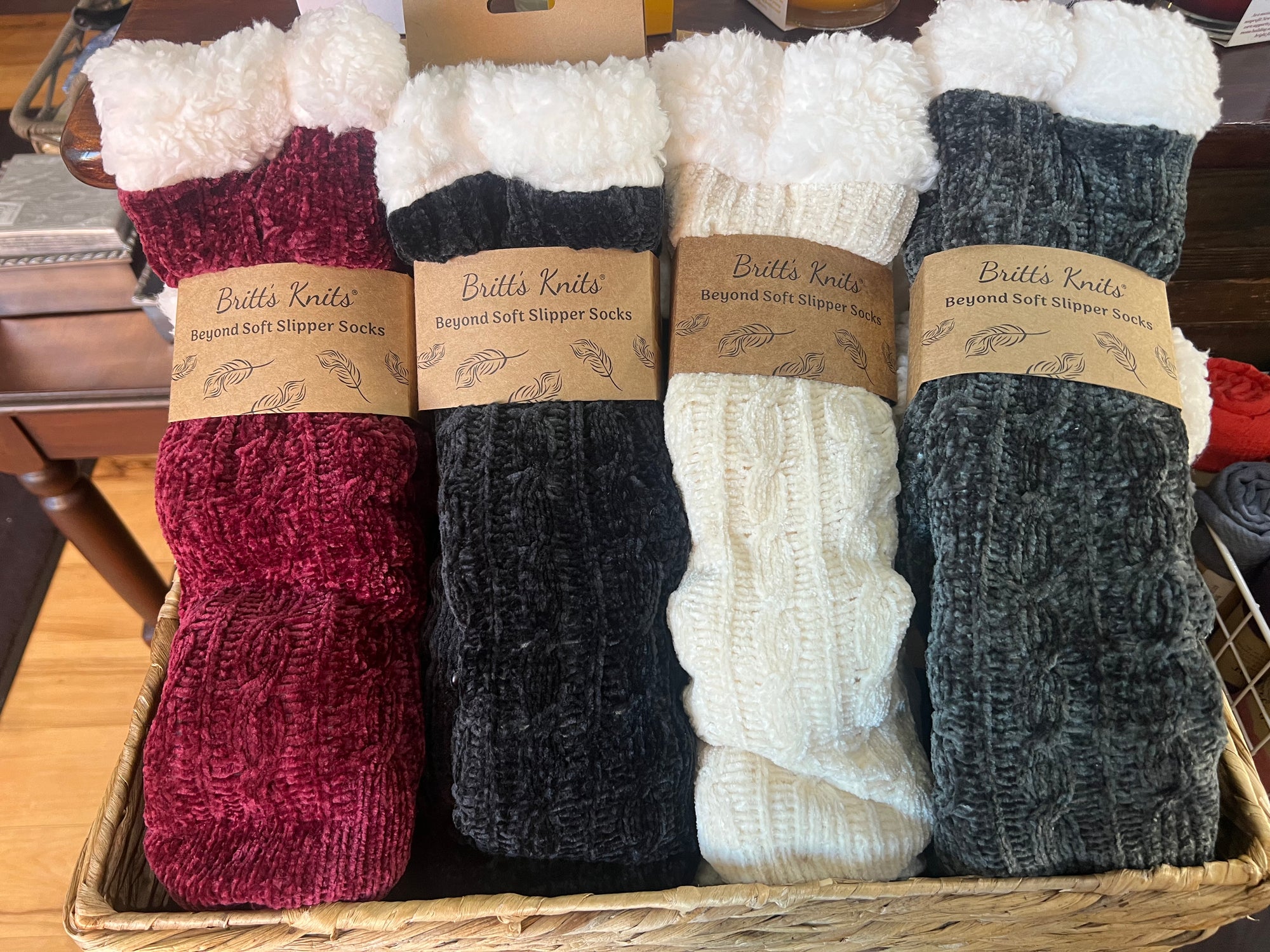 A basket holds four pairs of Beyond Soft slipper socks. The socks come in red, black, white, and gray, each with fluffy white cuffs. They feature non-slip soles and are packaged with brown labels, displayed on a dark wooden table.