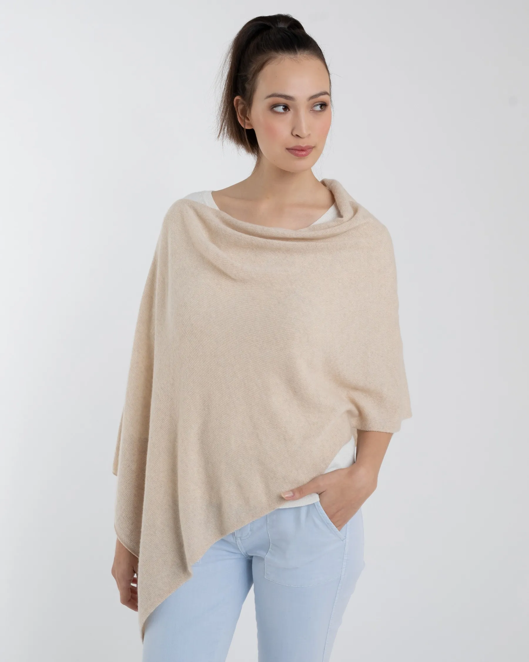 A person with long dark hair tied in a high ponytail is showcasing the Cashmere Poncho, styled over a white shirt and light blue jeans. They stand against a plain white background, gazing to the side with one hand in their pocket, exemplifying that this garment is truly one size fits all.