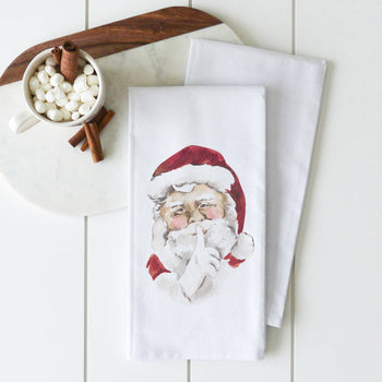 Part of our Tea Towels - Winter/Christmas collection, two white towels are elegantly displayed on a white table. The top towel features a handcrafted illustration of Santa Claus with a finger over his lips, signaling silence. Nearby, a cup brimming with marshmallows and cinnamon sticks rests on a marble cutting board.