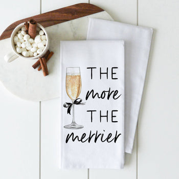A white tea towel from our Winter/Christmas collection features a printed champagne glass adorned with a black ribbon and the text "The more the merrier." It is displayed on a white wooden surface next to a small dish of marshmallows and cinnamon sticks on a marble board.