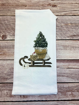 A white Tea Towel from the Winter/Christmas collection showcases an embroidered wooden sled carrying a handcrafted potted evergreen tree dusted with snow, surrounded by small wrapped gifts in festive colors on a rustic wooden surface.