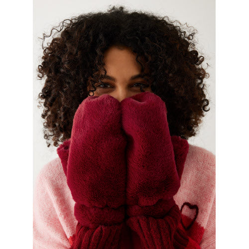 A person with curly hair playfully or shyly covers their face with Cozy Cabin Faux Fur Mittens in a plush red, offering only a glimpse of their eyes against the plain, light background. They sport a pink sweater decorated with a small red heart on the chest, while the added faux fur enhances the warmth of the scene.
