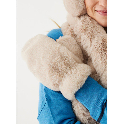 Wearing a vibrant blue sweatshirt, an individual hugs themselves in comfort while donning the Cozy Cabin Faux Fur Mittens in beige. A matching faux-fur scarf or hood partially conceals their face, with some hair peeking out. The backdrop is simple and white.