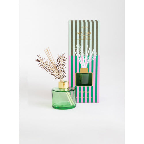 The Holiday Reed Diffuser, housed in a green glass bottle with a golden cap and adorned with leaf-shaped decorative reed sticks, emits a piney scent reminiscent of coastal pines. It is accompanied by a matching box featuring green, pink, and gray vertical stripes along with 