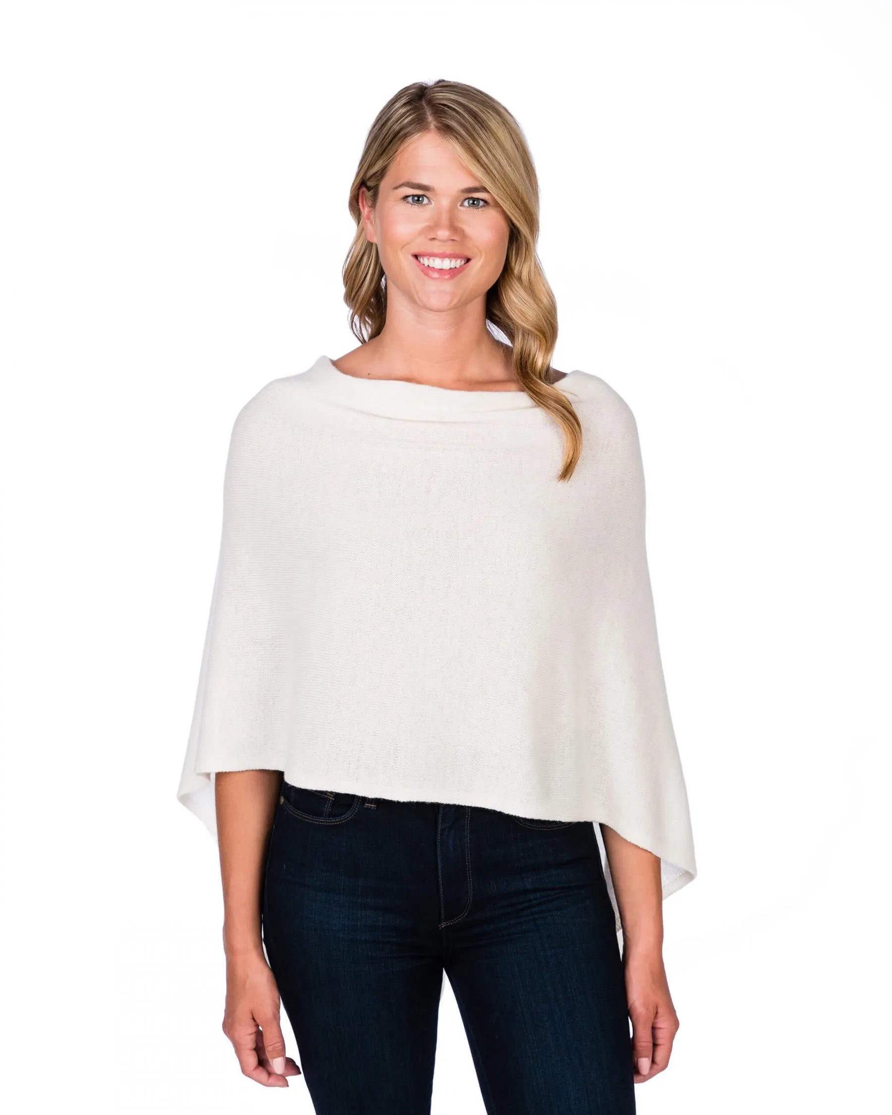 A smiling woman with long, wavy blonde hair is wearing the Cashmere Poncho in white and dark jeans. She stands against a plain white backdrop, facing forward. The poncho drapes elegantly over her shoulders, creating a chic dress topper that provides a sophisticated yet casual appearance.