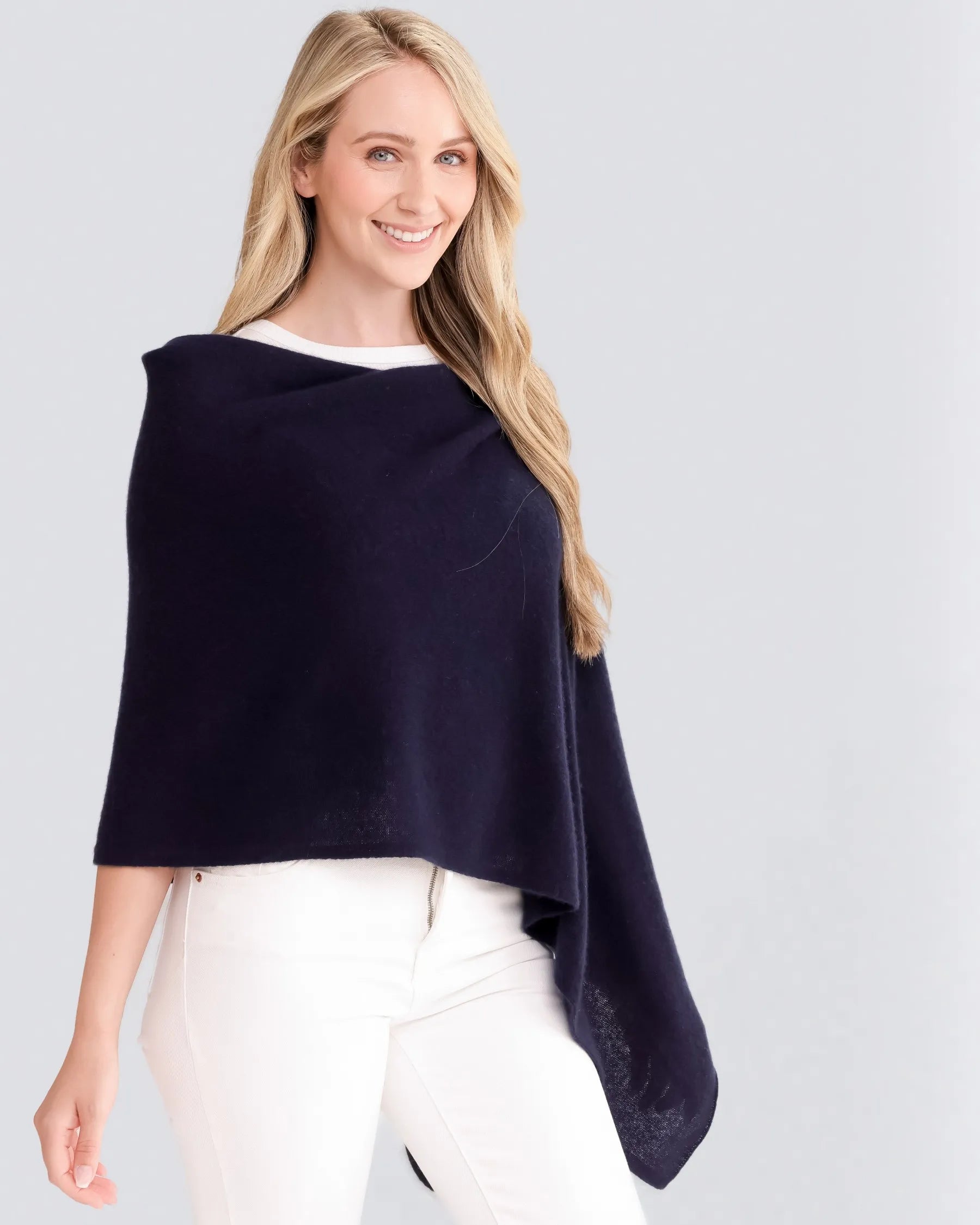A person with long blonde hair smiles as they pose against a plain light gray background, wearing the luxurious Cashmere Poncho in navy blue. The poncho drapes elegantly over their white top and white pants, creating a striking contrast. With its cozy, one-size-fits-all design, the person extends an arm slightly to the side to showcase this stylish dress topper.