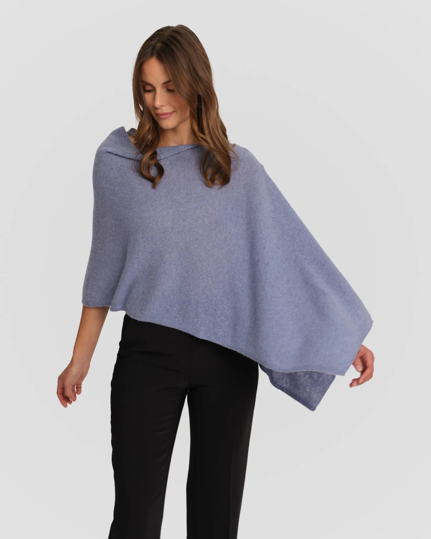A person with long brown hair is dressed in a light blue, one-size Cashmere Poncho and black pants. They are posing against a plain white background, gazing gently downward and to the side, with one arm extended gracefully. The elegant Cashmere Poncho drapes softly, forming gentle folds.