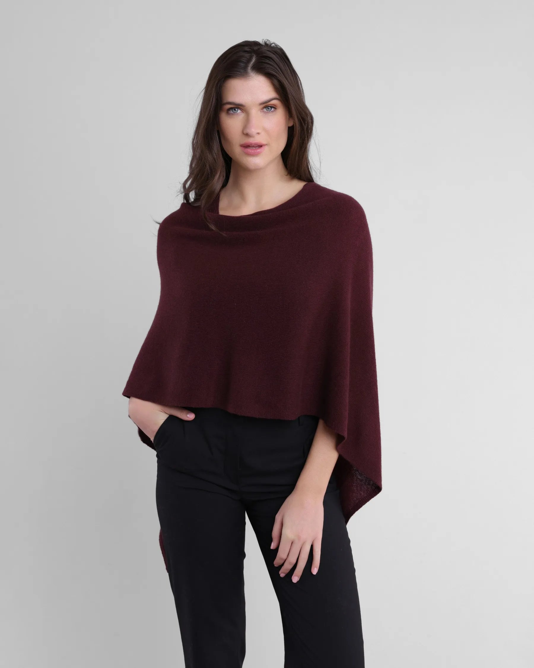 A woman with long brown hair wears a One Size burgundy Cashmere Poncho over a black outfit. This piece adds elegance as she stands against a neutral background, her left hand in her pocket and her right arm by her side, looking directly at the camera with a slight smile.