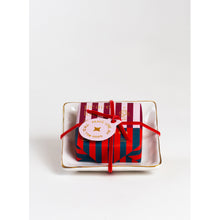 Load image into Gallery viewer, A small rectangular dish with gold trim holds a Holiday Soap in a Dish, wrapped in red and blue striped paper and tied with a red ribbon, exuding holiday cheer. A round tag attached reads &quot;PEACE&quot; and &quot;GOOD WILL,&quot; set against a plain white background reminiscent of a serene winter&#39;s day.
