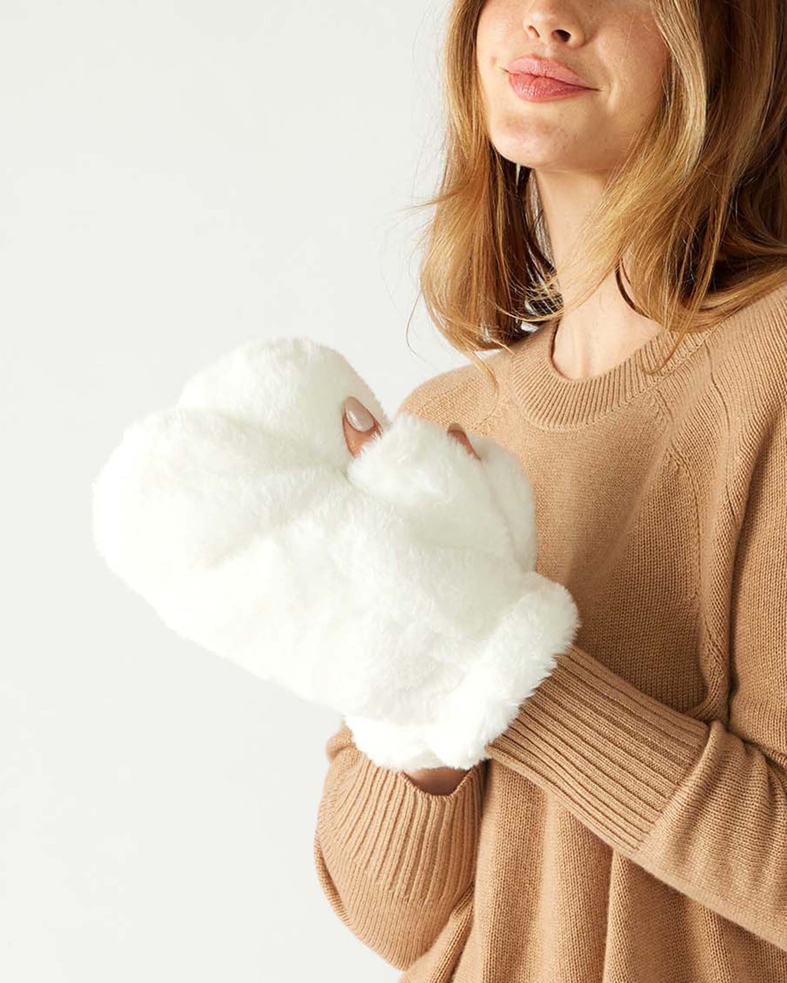 A person with shoulder-length hair in a beige sweater holds a pair of Cozy Cabin faux fur mittens. Against a plain, light-colored background that softly contrasts the cozy textures, they appear to be smiling gently.