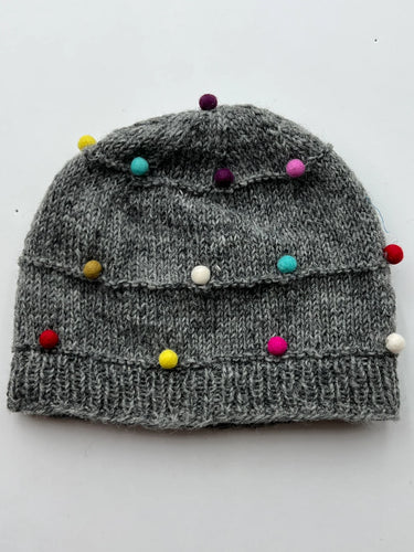 Introducing the Pom Pom Wool Hat—a gray knitted beanie made from soft sheep wool. This hat is adorned with vibrant pom-poms in yellow, blue, purple, pink, and white, bringing a playful charm to its neutral base. To ensure lasting comfort and quality, it's recommended to hand wash only. Displayed against a plain white background.