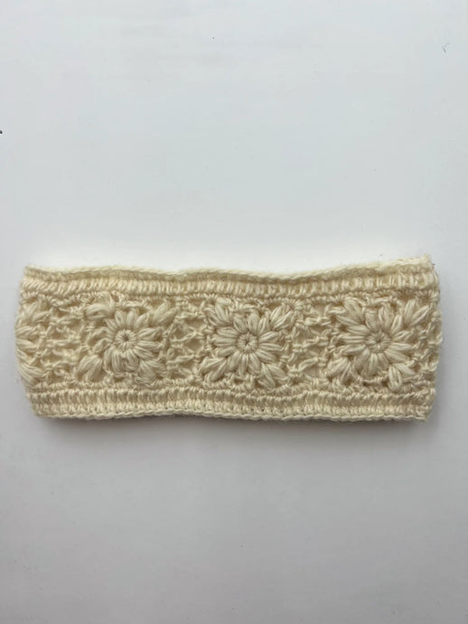 Introducing the Wool Knit Headband, a cream-colored accessory handmade in Nepal. This headband showcases an intricate floral pattern with delicately spaced flowers arranged in a row across the band. Fashioned from textured sheep wool, it beautifully highlights the detailed stitching of the floral design against its smooth white background.