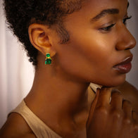A person with short curly hair is shown in profile, wearing a beige top and Gold Plated Sapphire Princess and Emerald Cut Drop Post Earrings that glimmer subtly. Their hand rests near their chin as they gaze thoughtfully off-camera, against a softly blurred background featuring white curtains.