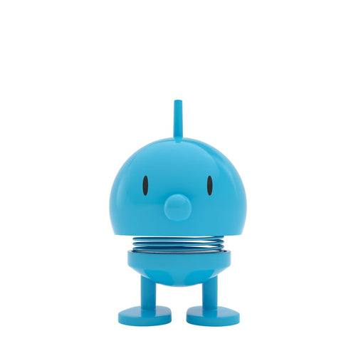 The Hoptimist Bumble is a small, round, blue toy figure with a smiling face and a spring body that captures the essence of Danish design history. It boasts a small nose, two black eyes, and a top fin, standing on two oval feet for a playful presence perfect for whimsical home decor.