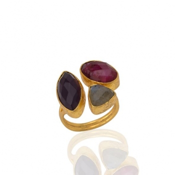 A gold ring with a distinct design showcases three large faceted gemstones: a deep purple Amethyst marquise, a vibrant red Ruby oval, and a triangular Labradorite stone in gray. These stones are beautifully set within a wide, open gold band for an elegant and artistic appearance.