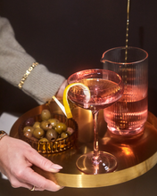 Load image into Gallery viewer, A hand holds one of The Lou Coupe Glasses from a set of two, featuring a pink hue and holding a craft cocktail garnished with a lemon twist. A matching pink pitcher and a bowl of green olives complement the scene. The person is dressed in a gray sweater adorned with bracelets. The dimly lit environment, enhanced by its mood-boosting colors, creates an inviting ambiance.
