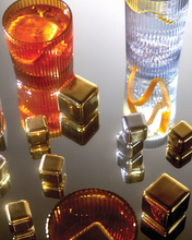 Load image into Gallery viewer, A reflective surface showcases two glasses: one featuring a textured design with an orange beverage and slices, and the other a clear glass adorned with a yellow spiraled peel. Around them lie the gleaming Ruby Cubes Cocktail Chillers in Gold – Set of 4, alongside transparent ice cubes. Warm lighting casts intricate reflections, accentuating their textures.
