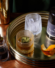 Load image into Gallery viewer, A set of The Jupiter Cocktail Chillers in Gold elegantly supports two textured glasses; one holds whiskey with a large freezable cube, while the other is filled with clear liquid and ice. Nearby sits a bottle of bitters, a dark drink labeled &quot;FASH,&quot; and an orange peel. The scene is arranged on a brown surface against a dark green backdrop.
