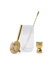 Load image into Gallery viewer, The Renley Bar Tool Set features a clear ribbed glass that holds a gold spiral cocktail stirrer, complemented by a gold cocktail strainer with a coiled spring resting against it. To the right, an elegant gold-plated stainless steel jigger stands, all beautifully arranged on a white background.
