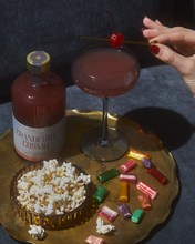 Load image into Gallery viewer, A brass tray presents a bottle labeled &quot;Cranberry Cosmo&quot; next to a cocktail glass filled and garnished with a cherry, skillfully placed on The Owen Cocktail Picks in Gold. Nearby, a small bowl of popcorn and vibrant candies beckon for snacking. A hand with red nails reaches for the enticing garnish against the gray fabric background.
