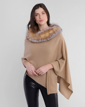 Load image into Gallery viewer, A woman with long dark hair is wearing the trendy Cashmere Fox Trim Poncho, showcasing its beige color and brown and gray faux fur collar. She pairs it with black leather pants, clasping her hands in front as she stands against a plain white background.
