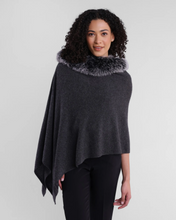 Load image into Gallery viewer, A person with curly hair is wearing a Cashmere Fox Trim Poncho in dark gray, featuring a luxurious fox fur collar. This stylish piece drapes asymmetrically over black pants, set against a plain white wall that highlights the fashion-forward clothing and style.
