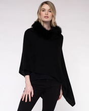 Load image into Gallery viewer, Standing against a light gray background, a person wears the Cashmere Fox Trim Poncho in black, featuring a luxurious fluffy black trim reminiscent of fox fur around the neck. With long blonde hair and a neutral expression directed at the camera, they stylishly pair this trendy piece with black pants or leggings.
