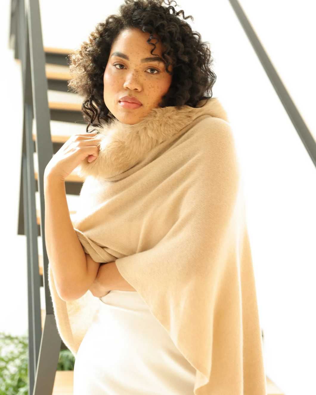 A person with curly hair is adorned in a Cashmere Fox Trim Poncho, standing in a well-lit space. They show a calm expression and are positioned in front of a staircase. The surroundings showcase modern, minimalist design with blurred plants visible in the background.