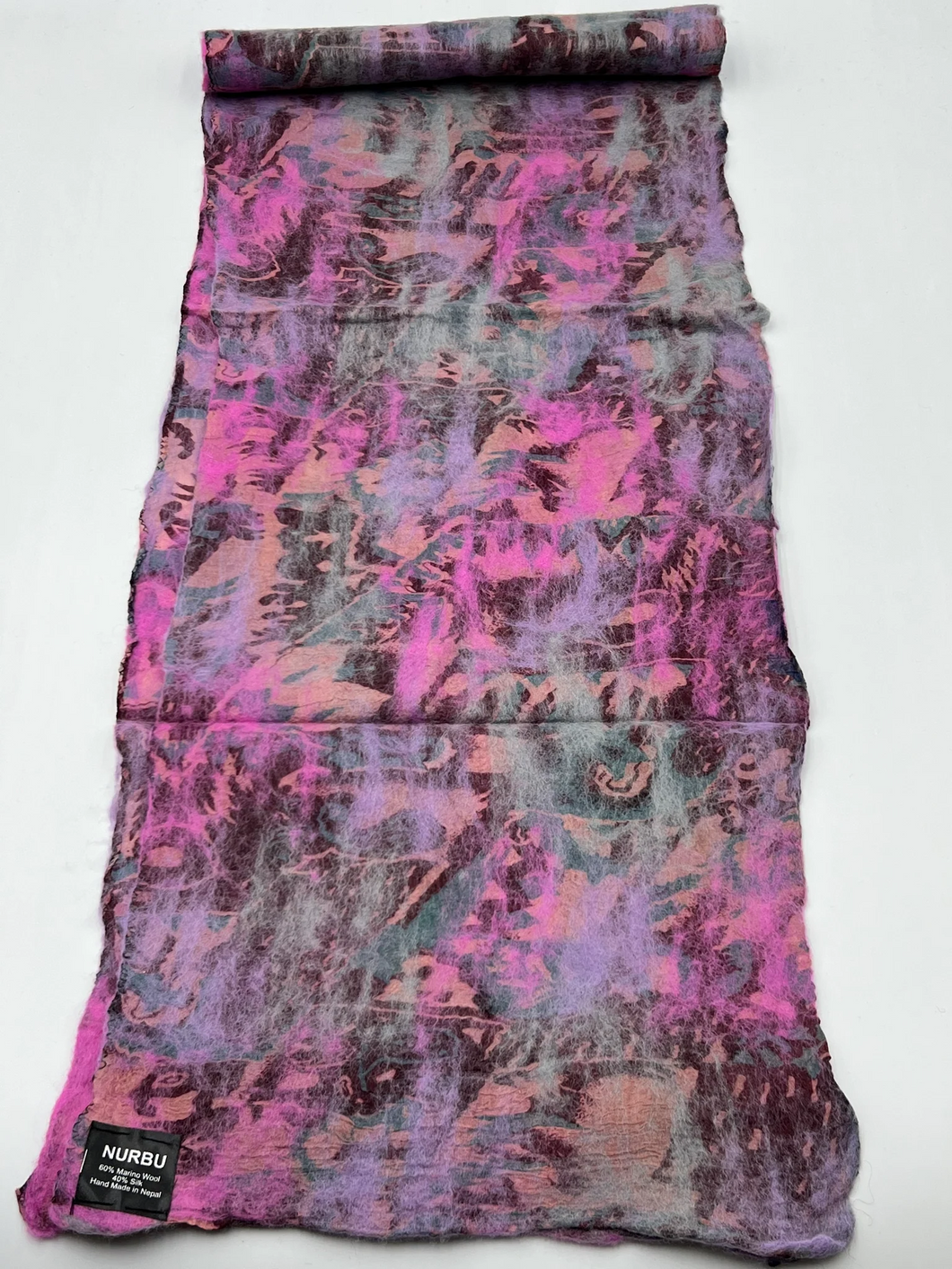 A vibrant, hand-rolled scarf made from merino wool and sari silk is spread out on a flat surface. The scarf showcases abstract patterns in pink, purple, and light blue hues. In the bottom left corner, a black label reads, 