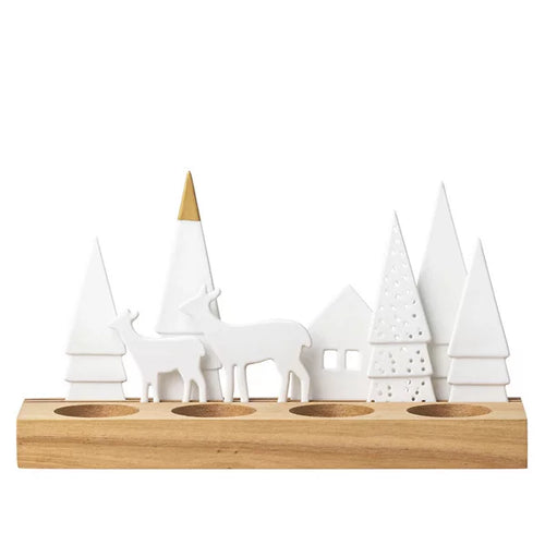 The Holiday Tealight Holder - Forest is a rustic cabin accessory featuring a wooden base adorned with a winter scene. It includes white porcelain figures of deer and triangular trees, some adorned with cut-out patterns. A small house stands prominently within the display, and the holder has four round recesses for candles, making it an ideal piece for holiday forest decor.