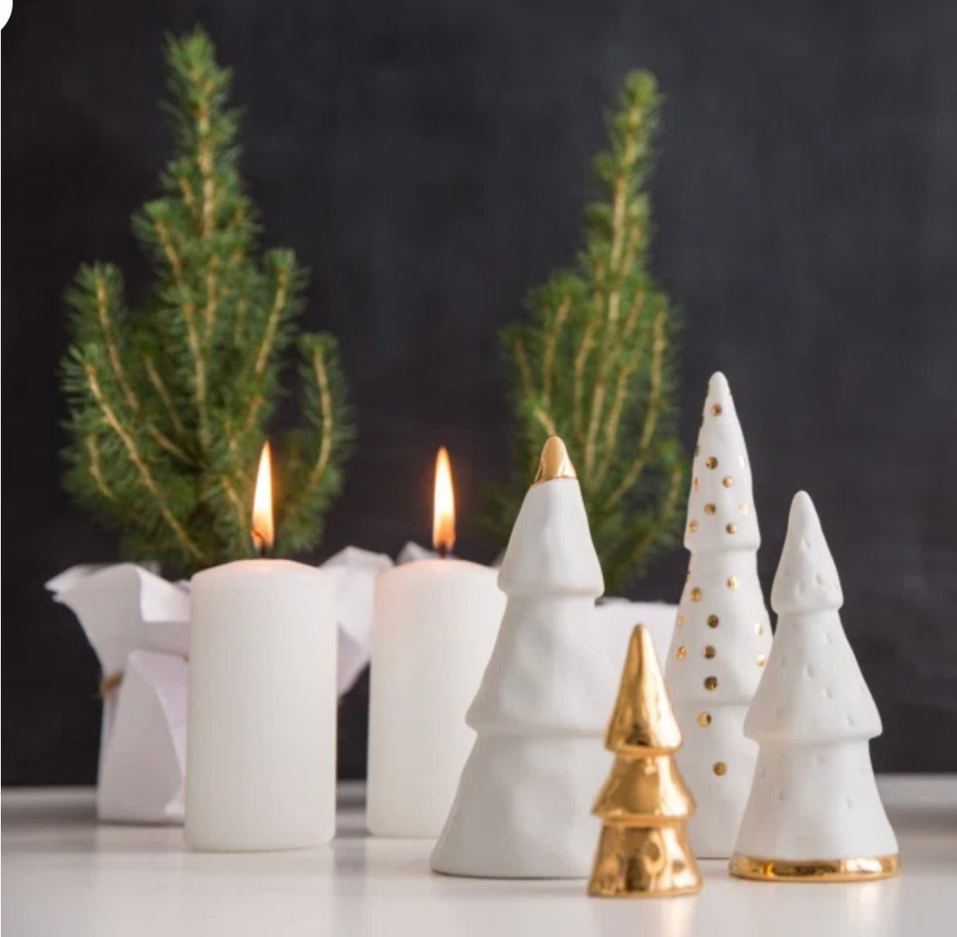 The Christmas Forest, featuring porcelain Christmas trees adorned with gold and white designs, is elegantly displayed on a white surface. In the background, two lit white candles wrapped in small pine branches stand against a dark backdrop, creating a festive and cozy atmosphere—ideal minimalist gifts for the season.