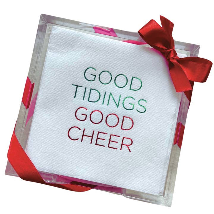 The Cocktail Napkin Acrylic Tray Set, featuring white disposable napkins printed with "GOOD TIDINGS GOOD CHEER" in festive green and red letters, is elegantly displayed in an acrylic tray. Ideal for holiday hosting, these napkins are adorned with a vibrant red ribbon tied into a charming bow on the right side.