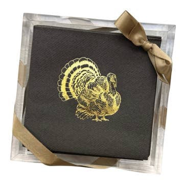 A stack of dark gray disposable napkins featuring a gold foil turkey design is neatly arranged in a clear, square holder from the "Cocktail Napkin Acrylic Tray Set - Holiday Themed." A gold ribbon tied in a bow adorns the corner, adding an elegant touch ideal for holiday hosting.