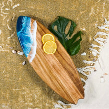 Load image into Gallery viewer, The Acacia Resin Surfboard, a wooden serving board shaped like a surfboard, rests on a sandy beach. Made from Acacia hardwood, this stylish cheese board features an eco-friendly resin section mimicking ocean waves on one end, with two lemon slices near the center. A large green leaf lies partly beneath, and scattered shells and fringe from a white cloth decorate the sand.
