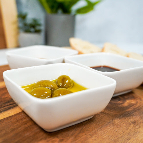 Close-up of three small Ceramic Dipping Bowls arranged in a row on a wooden surface. The foreground bowl contains green olives in oil, the middle bowl features a golden liquid, likely olive oil, and the background bowl holds a dark liquid that appears to be balsamic vinegar. Slices of bread and greenery are blurred in the background.