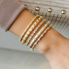 Load image into Gallery viewer, The scene focuses on a wrist elegantly decorated with five Marquis Pave Thin Hinge Bangles, each showcasing alternating patterns of cubic zirconia stones and intricate designs. The individual wears a ribbed gray sweater accented by decorative gold buttons on the sleeve, set against a softly blurred backdrop of neutral tones.
