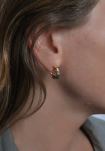 Load image into Gallery viewer, Close-up of a person’s ear adorned with the Bamboo Huggie Hoop Earrings, crafted in an 18k gold plated twisted design. This small, elegant piece complements the person&#39;s brown hair, with a few strands resting near the ear. The softly blurred background draws attention to these exquisite earrings.
