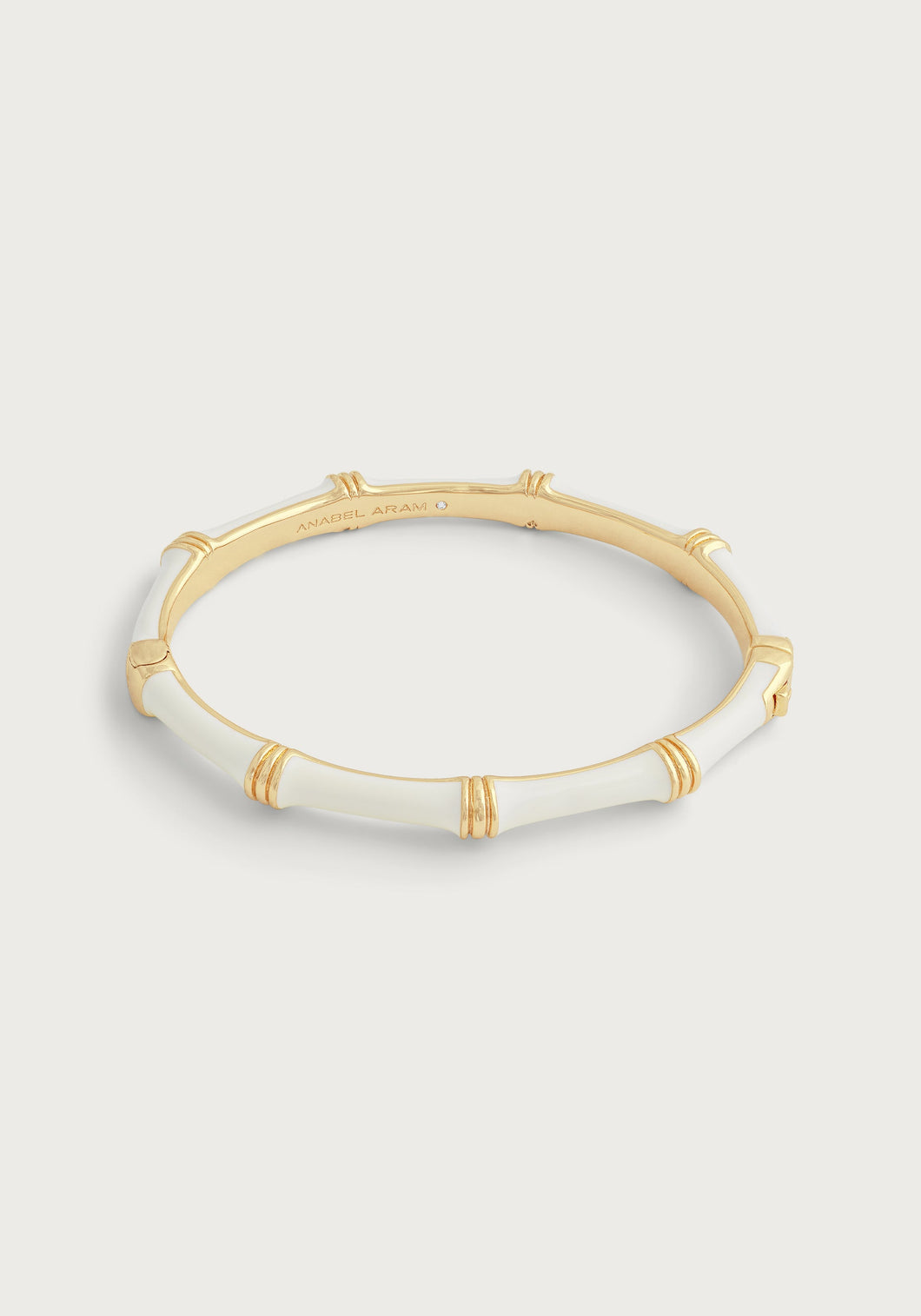 The Bamboo Enameled Hinge Bangle is an elegant bracelet crafted from 18k gold-dipped brass, featuring a bamboo-inspired design with smooth white enamel sections. Subtle engraved details near the joints and a polished finish enhance its sophistication, beautifully contrasted against a light grey background.