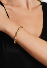 Load image into Gallery viewer, A person wearing the Bamboo Hinged Bangle, crafted from 18k gold-dipped brass, raises their arm to complement their black top. The sparkling crystals accentuate the bangle&#39;s unique textured elements, reminiscent of elegant bamboo stalks.
