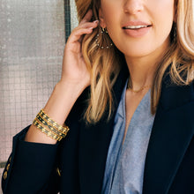 Load image into Gallery viewer, A woman touches her Signature Brooklyn in Bloom Open Hoop Earrings while wearing a navy blazer over a light blue blouse. Her wrist is adorned with multiple gold bracelets, and she wears a delicate gold necklace. The background features a textured glass surface, and her lips are relaxed in a soft smile.
