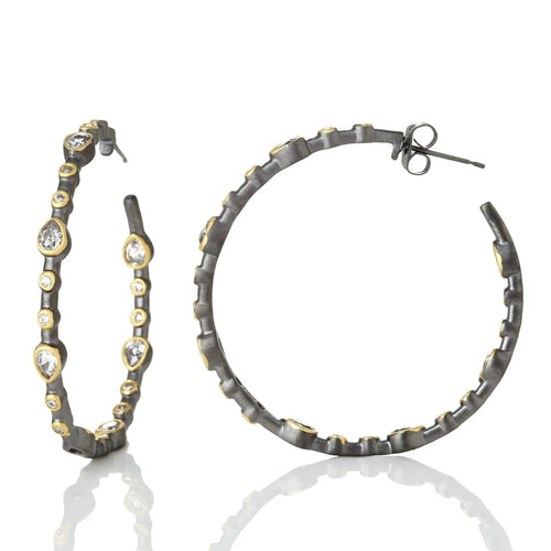 The Best Seller Hoops are showcased on a reflective surface, featuring two metallic hoop earrings embellished with oval and round gemstones of varying sizes in a prong setting along the outer edge, providing a striking contrast against the dark metal. The surface mirrors their intricate design and glimmering stones.