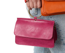 Load image into Gallery viewer, A person holds the vibrant pink Aria Ring Bag, crafted from vegan leather with a gold circular handle and flap design. They also carry an orange crossbody bag, paired with light blue jeans. The white background highlights the bags&#39; striking colors.
