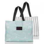 Two stylish Aloha Holo Holo Reversible Beach Totes: The front one is light blue with a subtle white pattern, black handles, and a black rectangular label. Behind it is a white tote with thin black stripes and matching handles. Both are on a plain background.