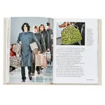 An open "Little Book of Louis Vuitton" reveals a luxury fashion runway scene on the left page, featuring models in dark attire with monogrammed bags. The right page displays text next to a close-up of a green patterned handbag, with the white background emphasizing the elegance of Louis Vuitton's design.
