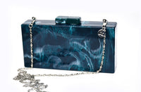 Load image into Gallery viewer, The Large Resin Clutch features a marbled dark teal and black finish with a sleek rectangular clasp. It includes a silver chain strap attached to round metal loops, elegantly draped for an artistic touch.
