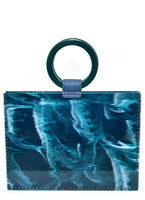 Load image into Gallery viewer, The Resin Valet Bag boasts a rectangular design with a teal and dark blue abstract pattern, featuring a circular dark green leather handle connected by a small blue strap. Its glossy marbled finish and sturdy vinyl bottom create an artistic and modern aesthetic.
